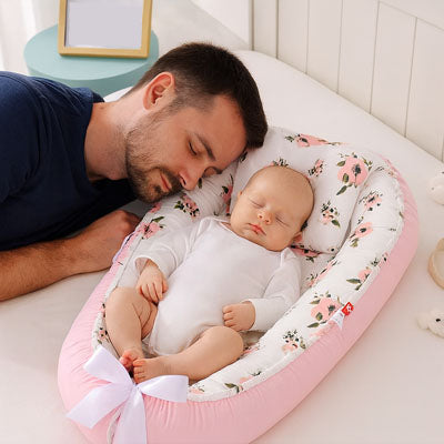 Père et bébé dormant paisiblement côte à côte dans un réducteur de lit bébé rose à motifs floraux, favorisant le cododo sécurisé.